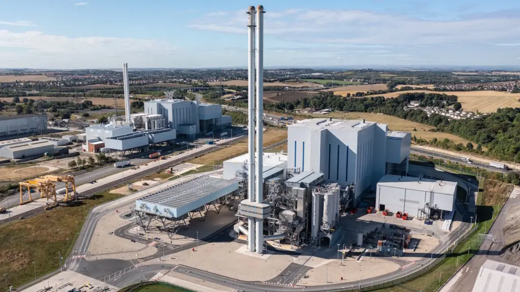 Photograph of an Energy from Waste (EfW) plant featuring a modern industrial building with a tall chimney emitting minimal steam. The facility is surrounded by a clean, landscaped area with greenery, under a clear blue sky. The image highlights sustainable waste management processes that convert waste into renewable energy.